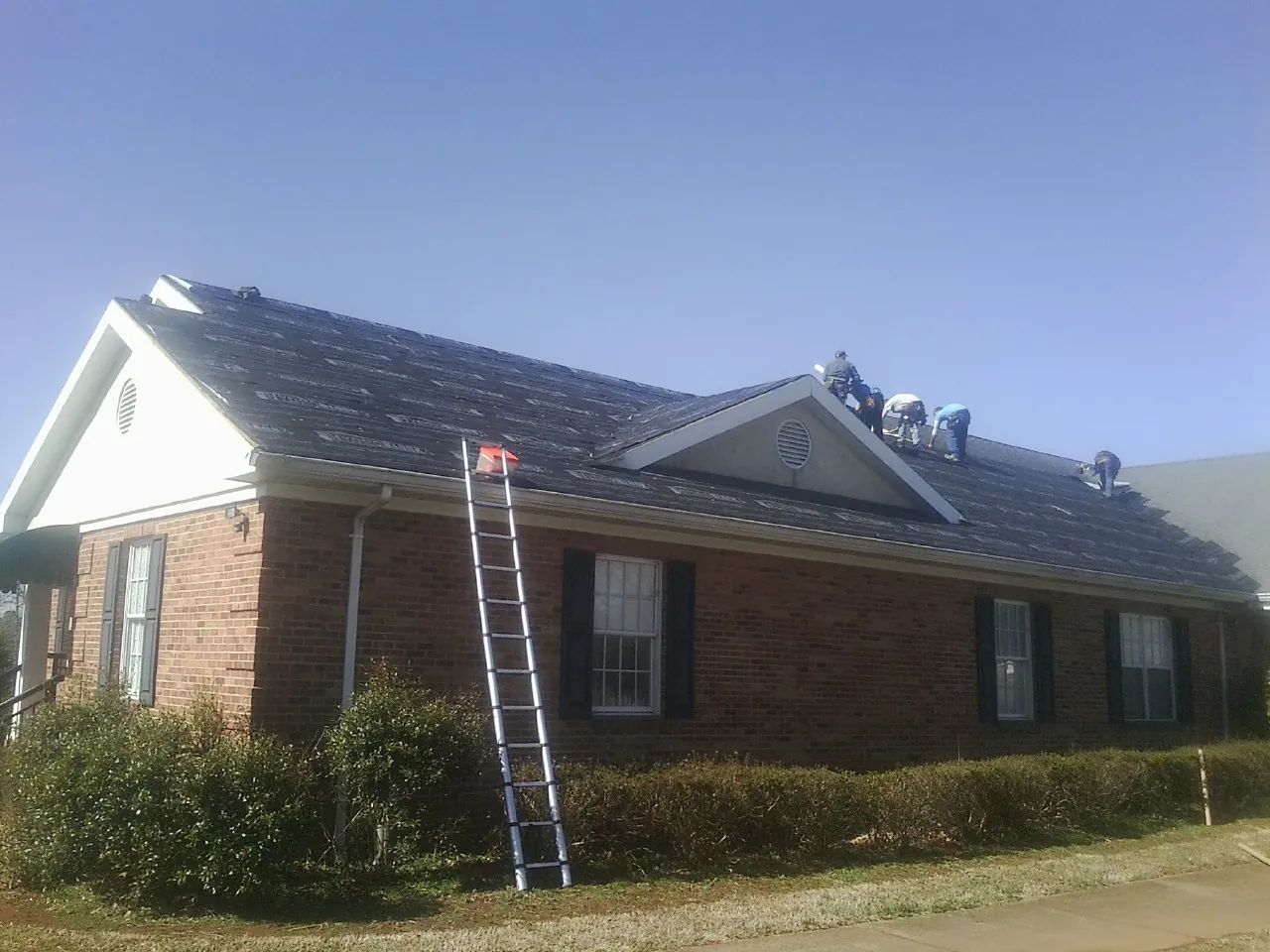 Crew tearing shingles off a brick ranch roof with a ladder set — Emergency Roof Repair work by our crew in Muir